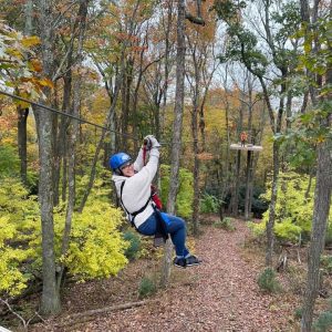 A person is zip-lining through a forested area with autumn foliage, wearing a blue helmet and harness, and smiling at the camera.