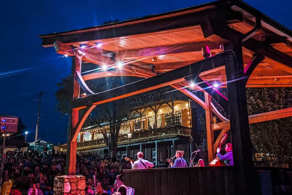 A stage setup with the band, Arcadian Wild, performing at night, surrounded by an audience. Colorful lights illuminate the scene, and a building with outdoor seating is visible in the background.