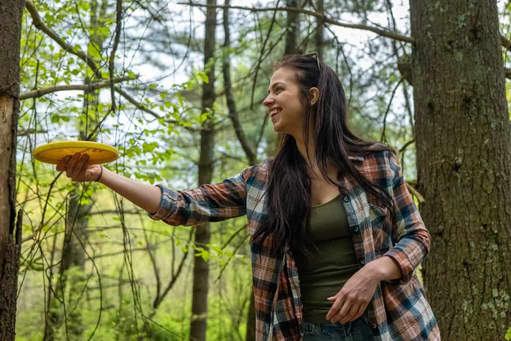 A woman with long, dark hair wearing a plaid shirt is throwing a yellow disc with a forest behind her.