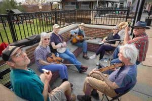 A group of seven musicians sitting together, playing various instruments like guitars and violins in a casual outdoor setting.