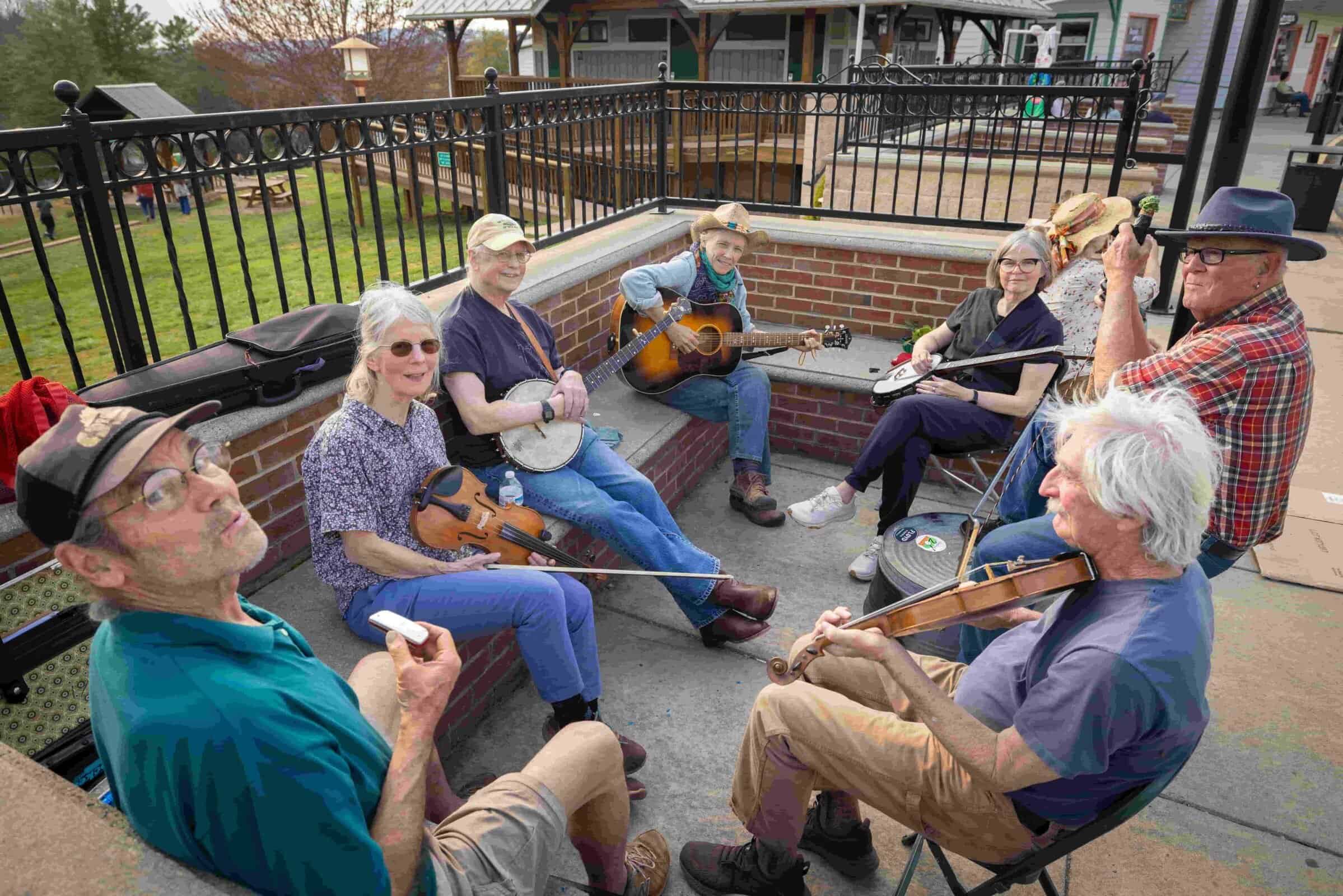 A group of seven musicians sitting together, playing various instruments like guitars and violins in a casual outdoor setting.