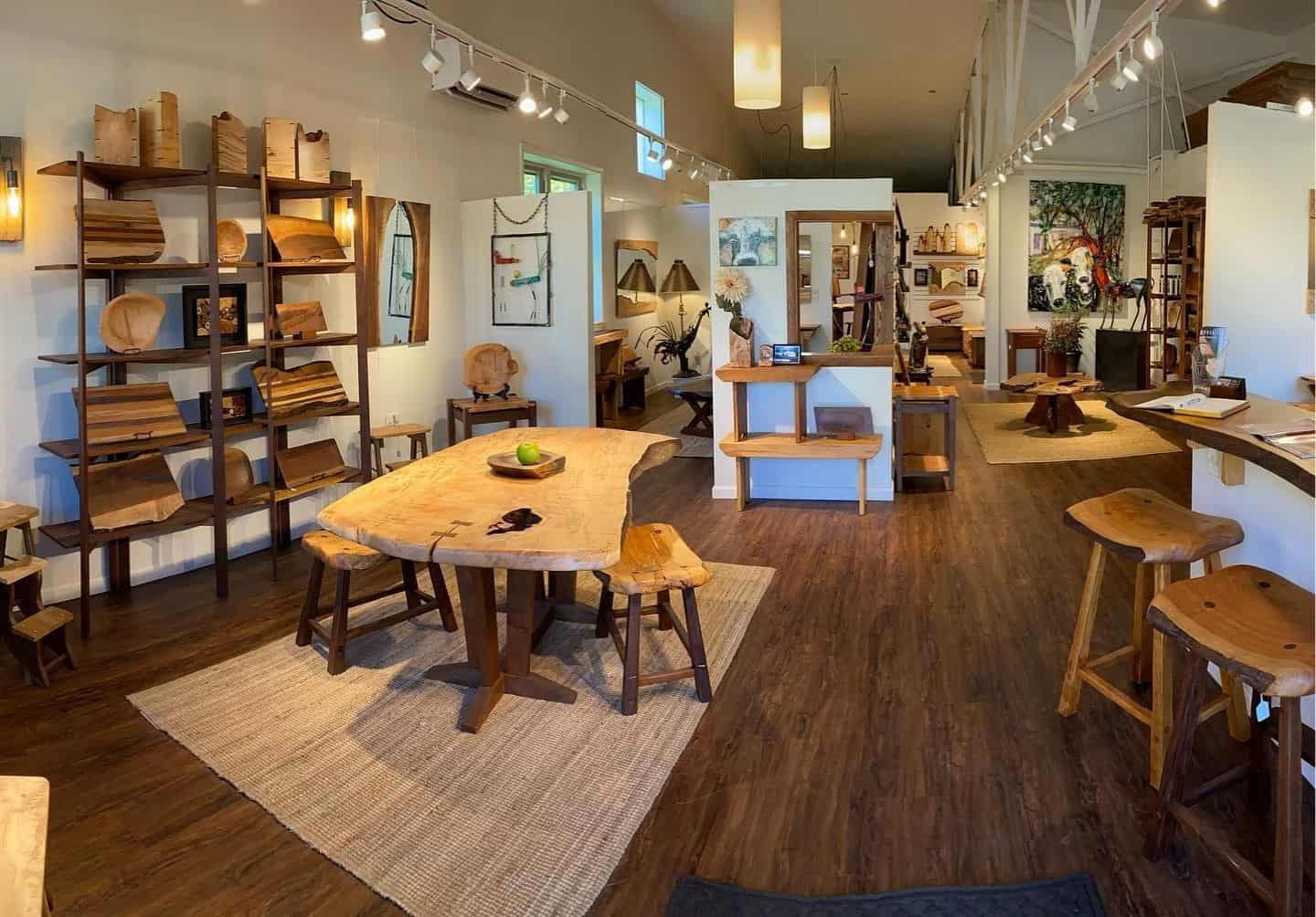 Inside view of the store, Phoenix Hardwood, showcasing a variety of handmade wooden furniture, including a large table, stools, shelves displaying wooden items, and decorative art pieces on the walls.