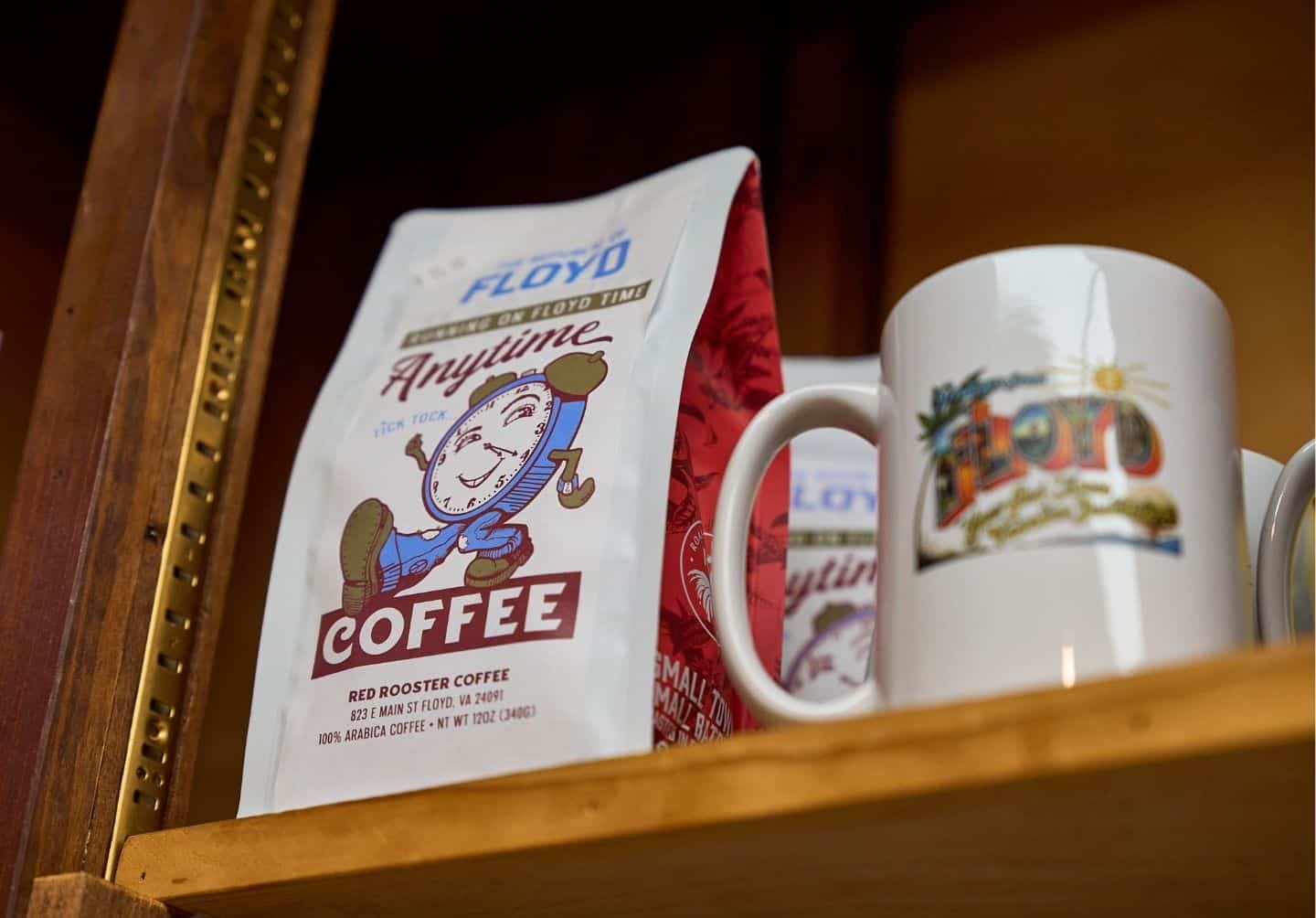 A bag of "Anytime Coffee" from Red Rooster Coffee and a white coffee mug with a colorful design, both displayed on a wooden shelf.