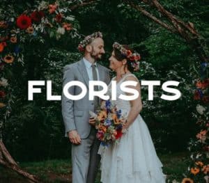 A couple stands together under a floral archway, with the bride wearing a white dress and holding a colorful bouquet, and the groom in a light gray suit.