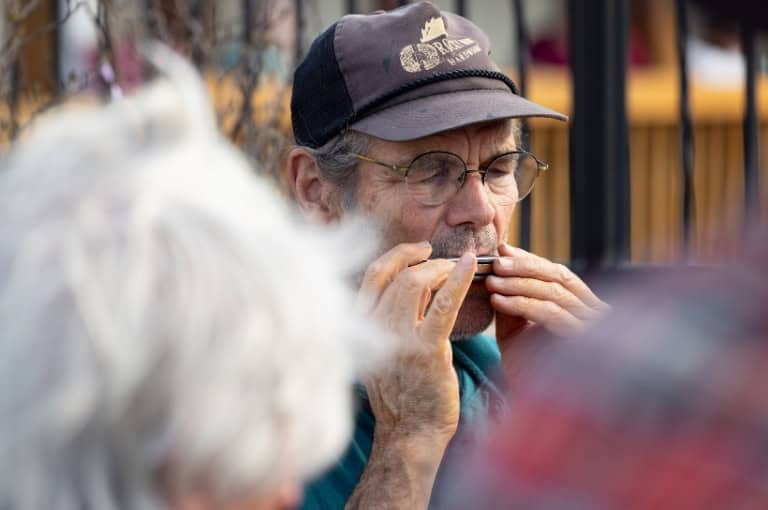A man playing a harmonica, wearing a cap and glasses, with blurred figures in the foreground.