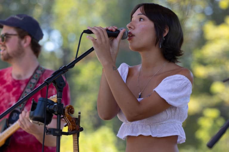 A woman in a white dress sings into a microphone, while a guitarist is partially visible in the background.