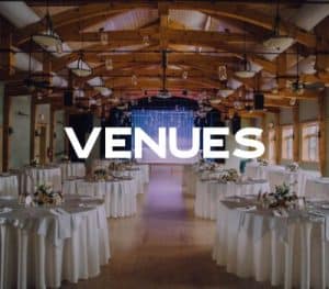 A beautifully set banquet hall with round tables adorned with white tablecloths and floral centerpieces, featuring a stage with draped lighting in the background.