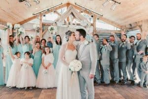 A bride and groom kiss at their wedding, surrounded by a joyful wedding party in light blue and gray attire, with guests holding flowers and celebrating.