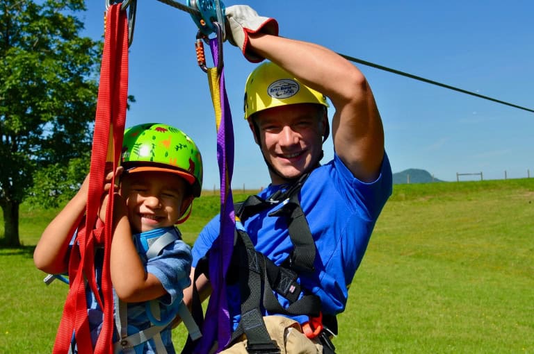A smiling adult and child are harnessed and ready for zip-lining, set against a green landscape. Both are wearing helmets.