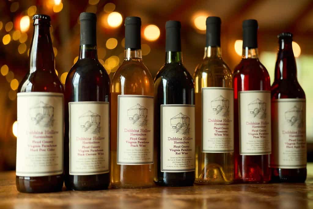A variety of bottles of wine and cider displayed on a wooden surface, with labels indicating different flavors and types, against a blurred background with warm lights.