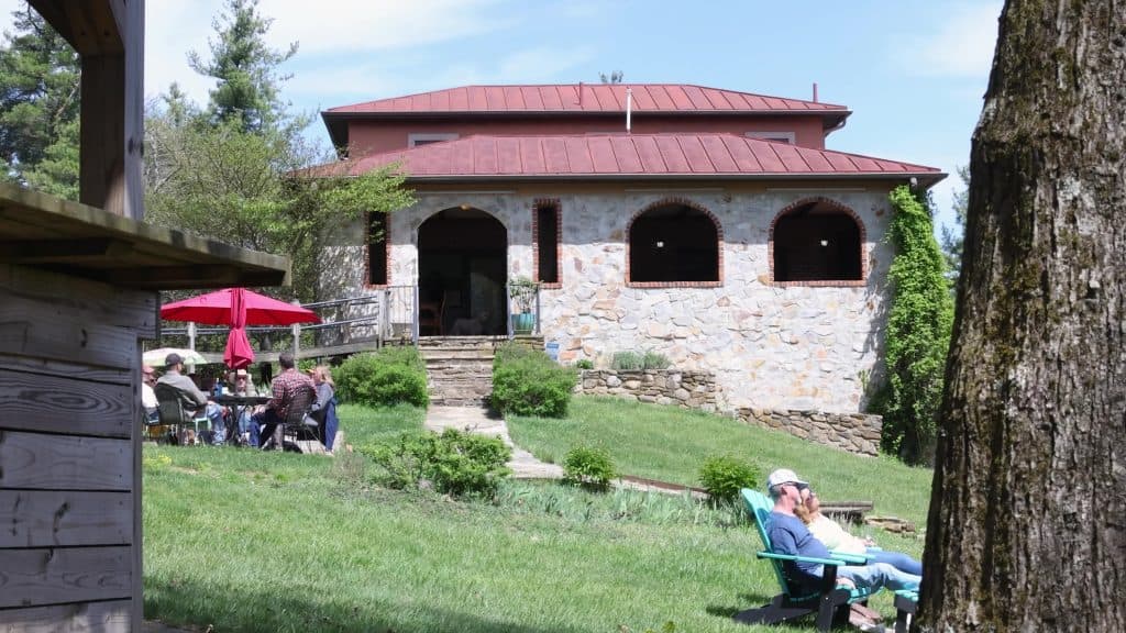 A stone and wood building with a red metal roof, surrounded by green grass. Several people are sitting around tables under umbrellas, while two individuals relax in lounge chairs in the foreground.