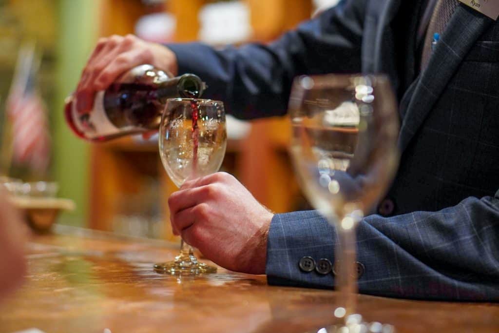 A person is pouring red wine into a glass at a bar, with another glass visible in the foreground.