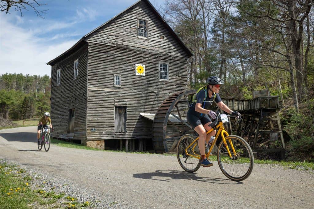 Two cyclists ride past a vintage wooden mill along a gravel path on a sunny day.