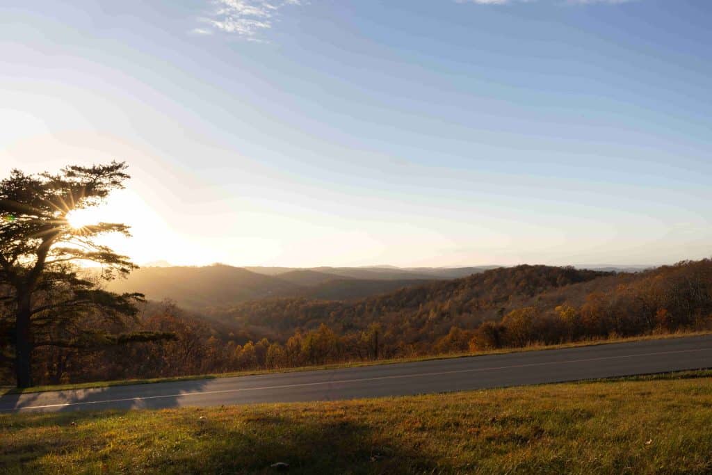 A scenic sunset from the Saddle Overlook on the Blue Ridge Parkway, featuring hills covered in autumn foliage, with a winding road in the foreground and a tree casting shadows.