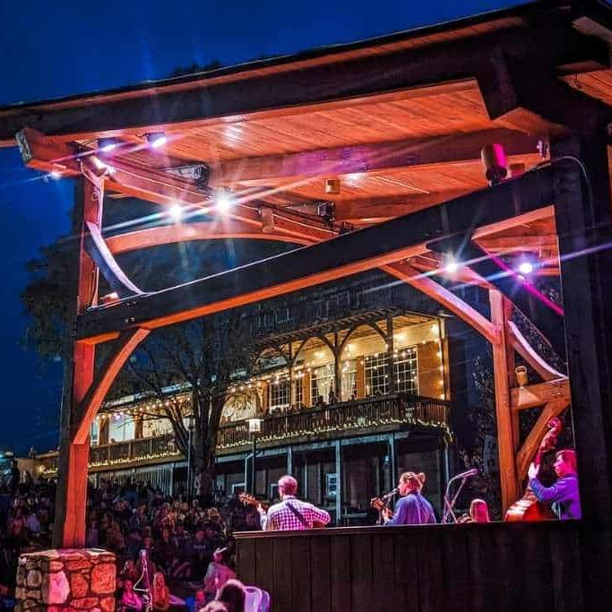 A stage setup with the band, Arcadian Wild, performing at night, surrounded by an audience. Colorful lights illuminate the scene, and a building with outdoor seating is visible in the background.