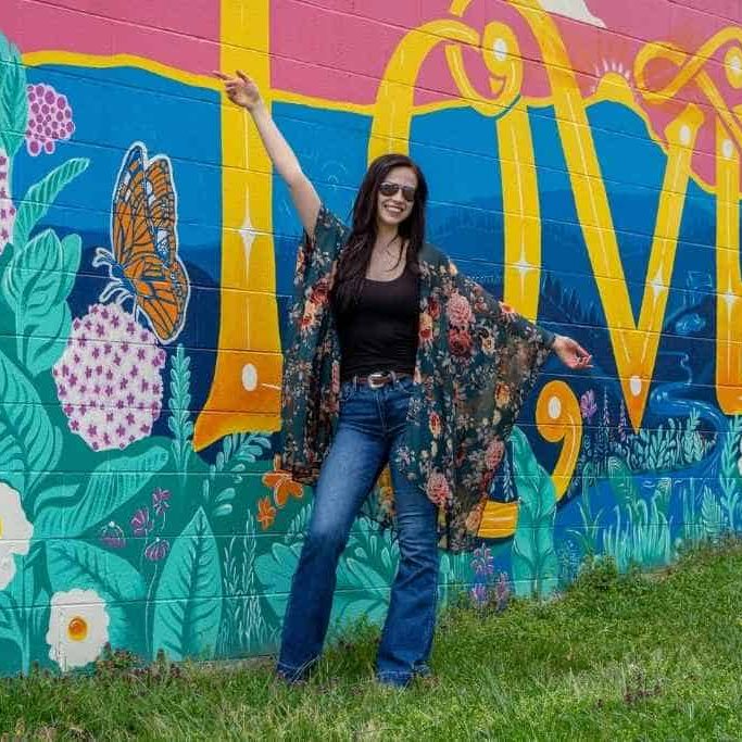 A woman in a floral kimono poses in front of a colorful mural featuring the word "LOVE" and vibrant flowers and butterflies.