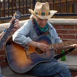 A musician wearing a straw hat and scarf is playing an acoustic guitar while sitting next to another person with a stringed instrument.