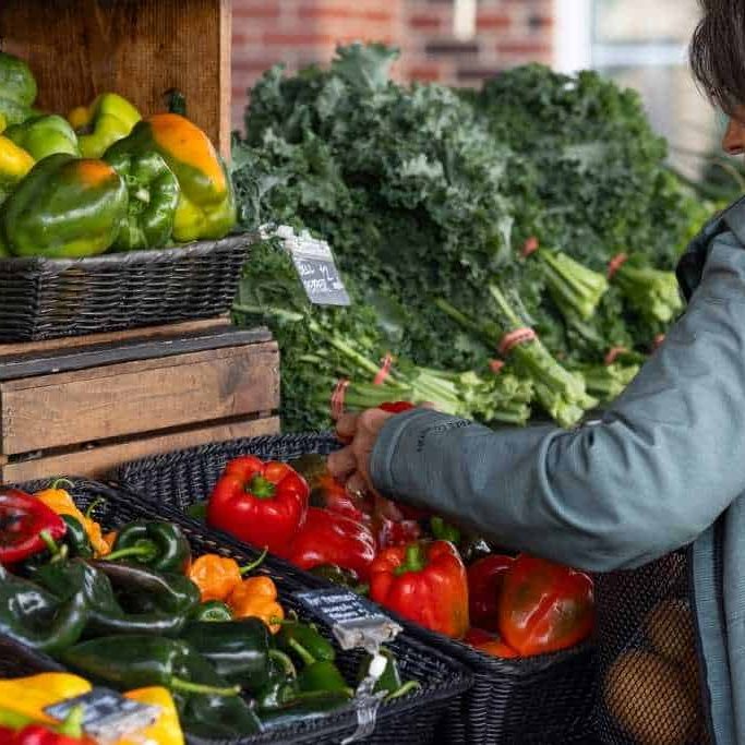 A person shopping for vegetables at a market, selecting red and green peppers, with baskets of various other vegetables in the background.
