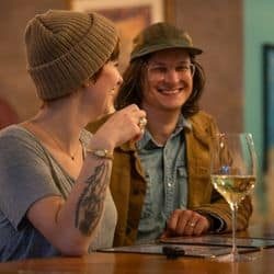 Two people sitting at a bar, smiling and chatting, with a glass of white wine in front of one of them.