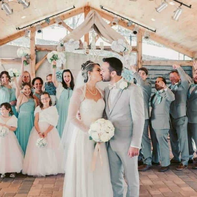 A bride and groom kiss at their wedding, surrounded by a joyful wedding party in light blue and gray attire, with guests holding flowers and celebrating.